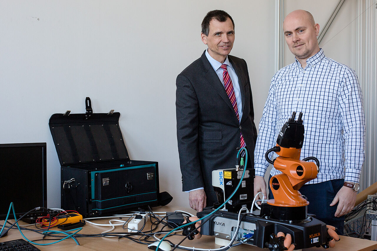 Ein Portrait von Prof. Jens Jäkel und Robert Thiel, vor ihnen steht ein Roboterarm auf dem Tisch.