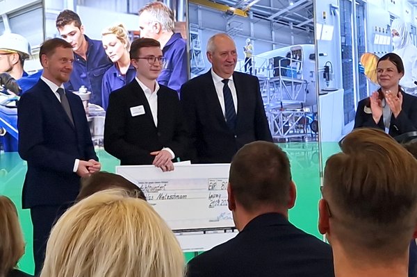 Gruppenfoto, Preisträger hält riesigen Scheck in der Hand, im Hintergrund Leinwand mit Bildern, im Vordergrund die Hinterköpfe der Zuschauerinnen zu sehen