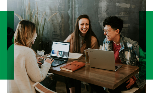 Drei Frauen sitzen zusammen am Tisch und reden miteinander. Vor ihnen stehen Laptops, Bücher und Getränke.