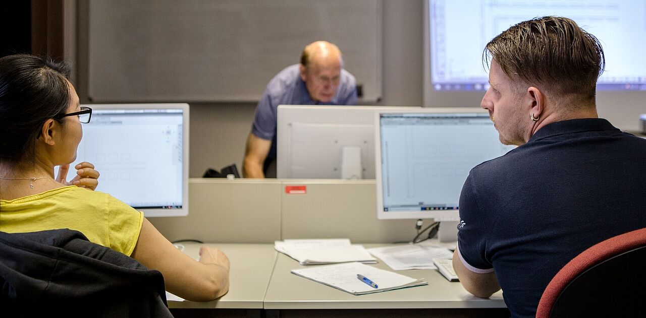 Zwei nebeneinander sitzende Lehrgangsteilnehmer eines PC-Kurses tauschen sich aus, während der Kursleiter im Hintergrund über seinen Dozentenrechner gebeugt steht.