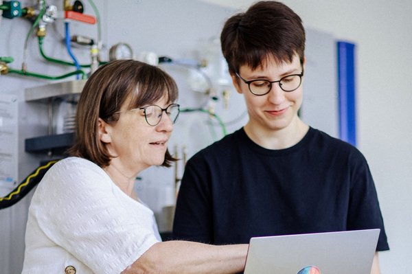 Technikbesprechung am Laptop Zwei Personen stehen vor einer technischen Wandinstallation. Eine zeigt auf den Laptop der anderen, beide tragen Brillen und wirken konzentriert.