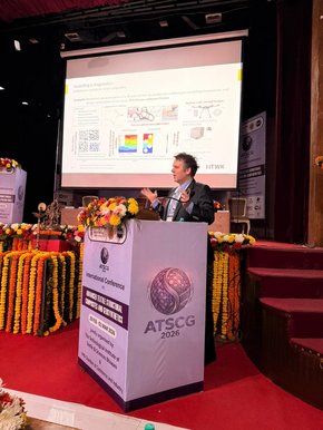 Prof. Robert Böhm steht auf einer Konferenzbühne und hält einen Vortrag. Im Hintergrund ist eine Präsentationsfolie zu sehen.