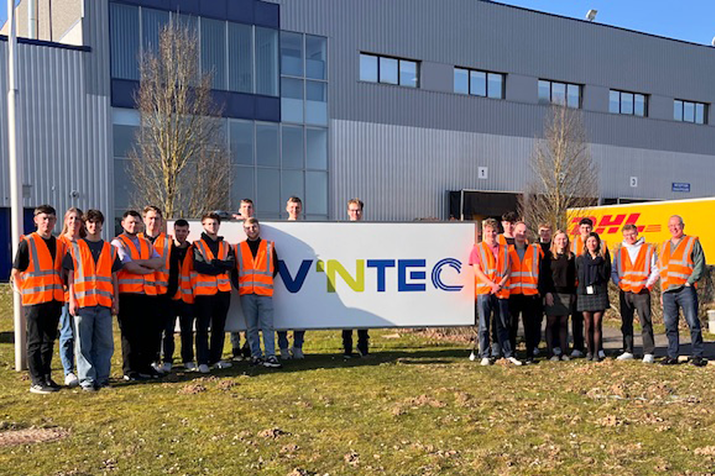 Gruppenfoto der Studierenden in Leuchtwesten auf dem Firmengelände, dahinter eine Fabrikhalle mit Firmennamen