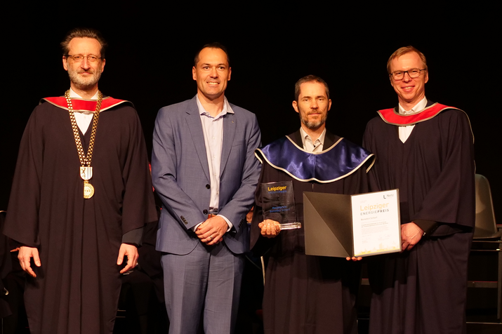 Gruppenfoto mit Rektor, Preisverleiher, Prof. Huhn und Dekan, Prof. Huhn hält Urkunde und gläsernen quadratischen Preis in der Hand