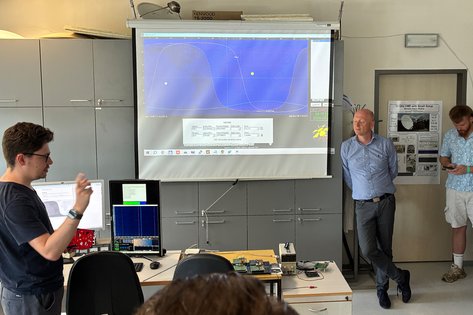 Labor ähnlich Seminarraum, BeamerLeinwand und diverse Technik im Raum, Person präsentiert auf Leinwand etwas, Studierende stehen herum