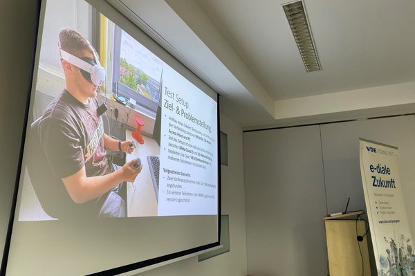 Test-Setup mit VR-Brille. (c) VDE Hochschulgruppe Leipzig Leinwand zeigt Präsenatation mit Bild einer Person mit VR-Brille am Schreibtisch sitzend