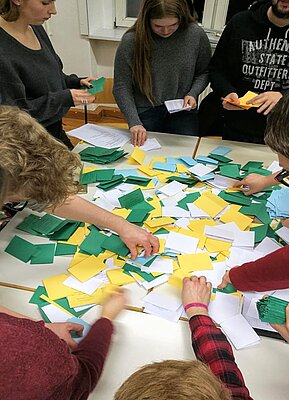 Eine Hand voll junger Menschen steht um einen Tisch herum und sortieren konzentriert Stimmzettel in vier verschiedenen Farben, die durchmischt auf dem Tisch liegen.