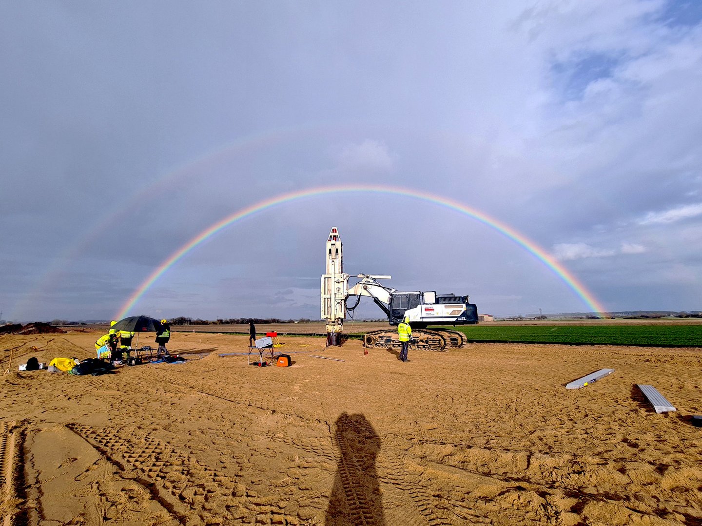 Impulsverdichtung auf der A14 ein großes Sandfeld, darauf steht eine weiße Maschine, dahinter sind 2 Regenbögen zu sehen