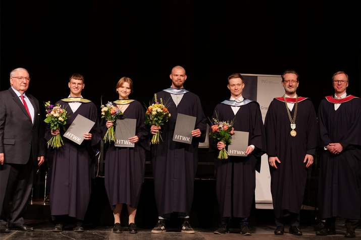 Gruppenfoto, alle stehen auf der Bühne des Werk 2, dunkler Hintergrund, Preisträgerin und Preisträger in Talar gekleidet haben Blumensträuße und Urkunden in der Hand, manche schauen in die Kamera