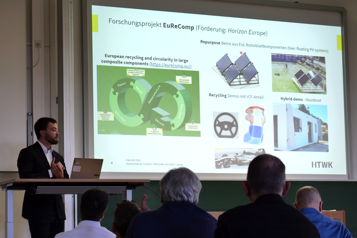 Dozent steht am Rednerpult, Powerpoint leuchtet an der Wand, davor Publikum von hinten zu sehen