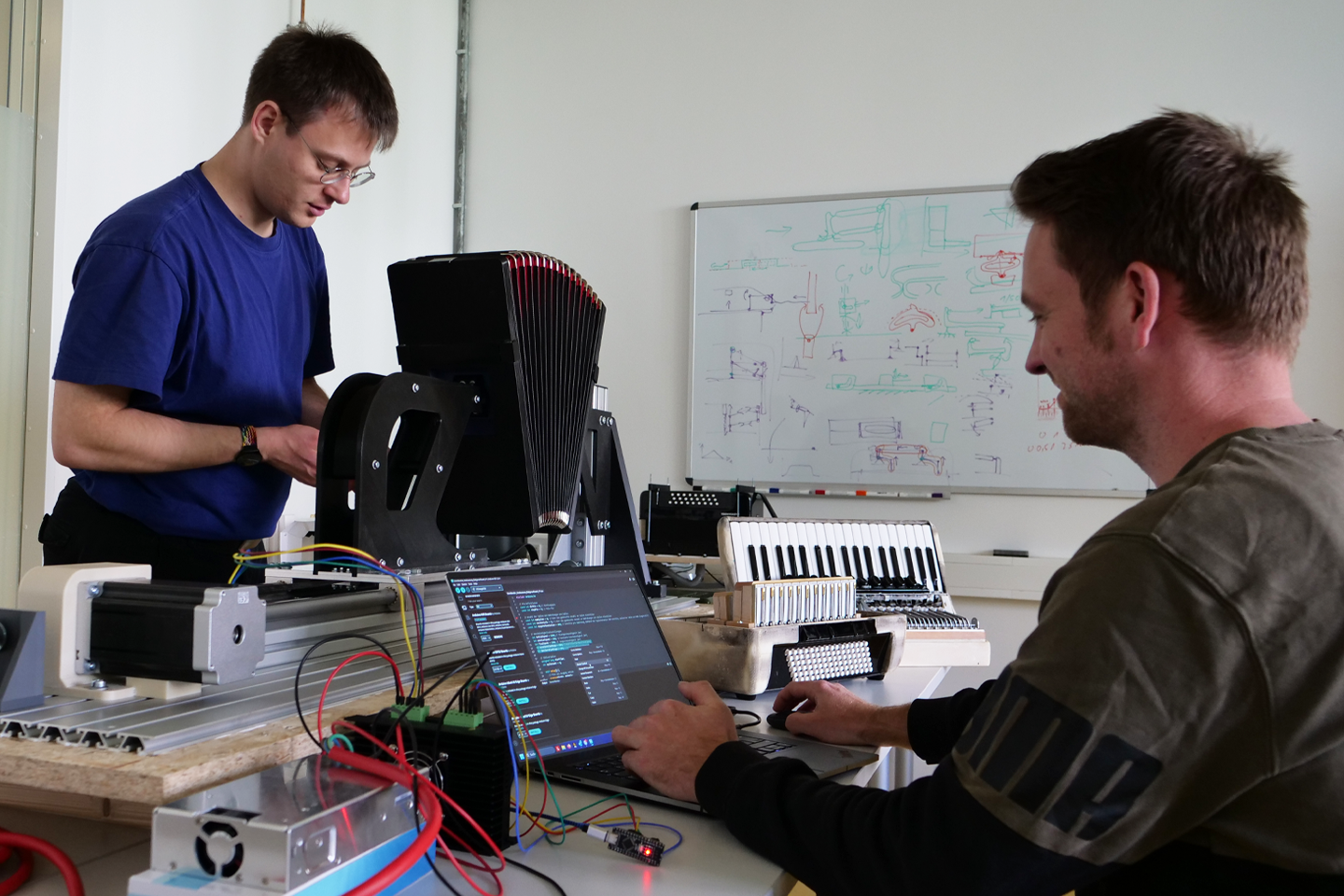 Wissenschaftliche Mitarbeiter arbeiten an einer Maschine mit Laptop und Accordeonprüfwerkzeug im Hintergrund ist ein auseinander gebautes Accordeon zu sehen