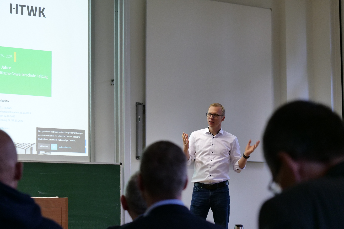Dean looks at the audience; next to him on the wall, part of the presentation can be seen (projected by a projector).