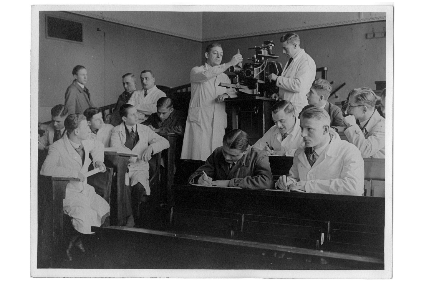 Black-and-white photo with students in white coats standing next to an old projector