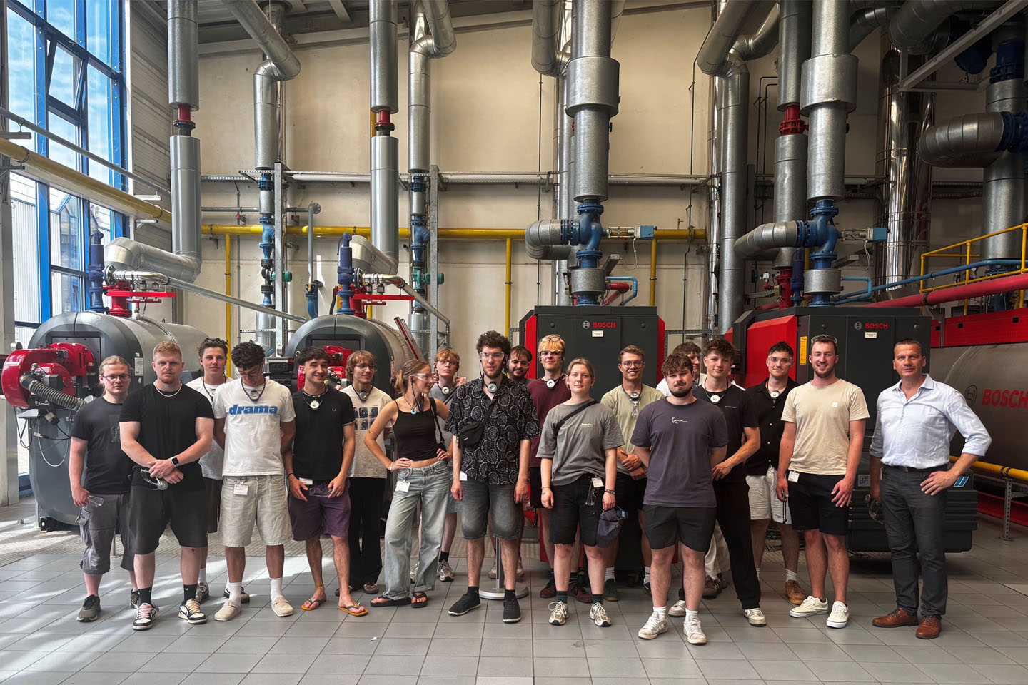 Seminargruppe steht in einer Werkshalle mit großen Röhren im Hintergrund, einige haben Sicherheitshelme auf - Gruppenfoto