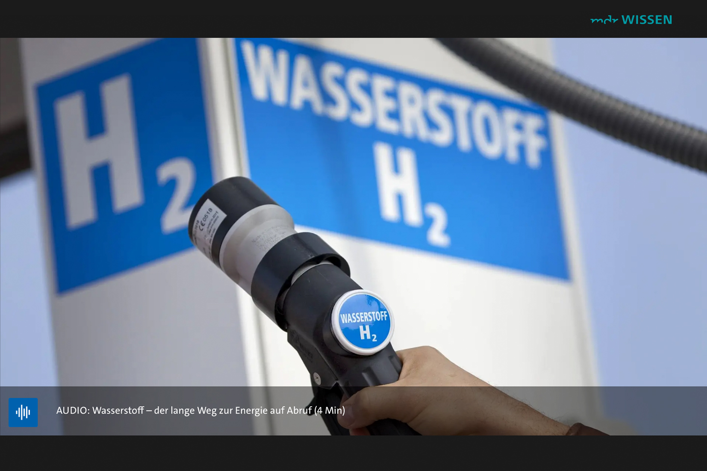 A charger similar to an electric vehicle charger with “H2” labeled on it for hydrogen; in the background, a large blue-and-white hydrogen display