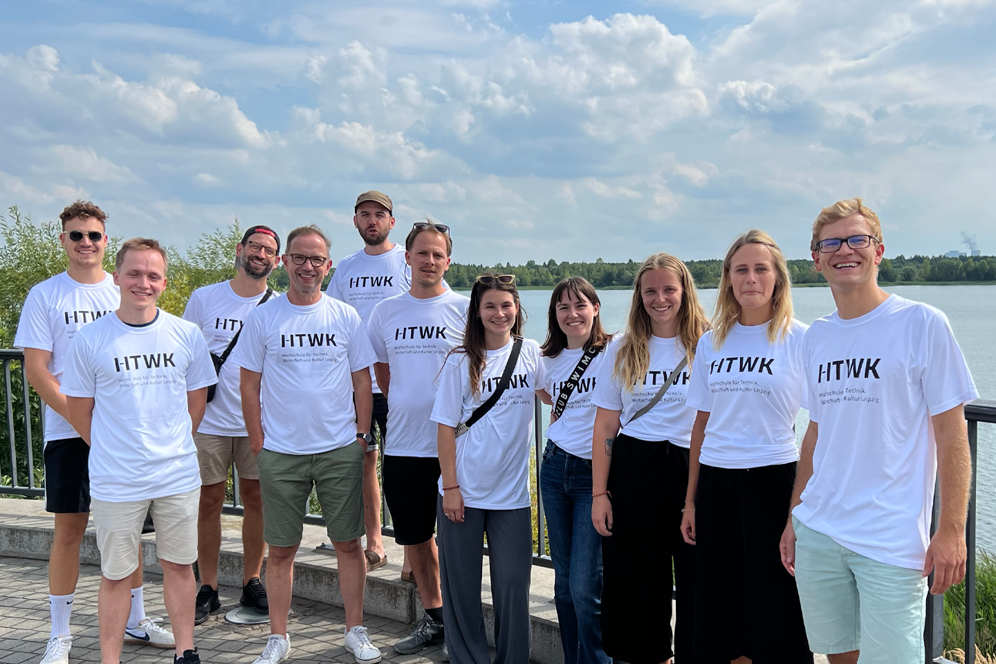 Group photo by the lake; everyone is wearing white shirts with the HTWK logo