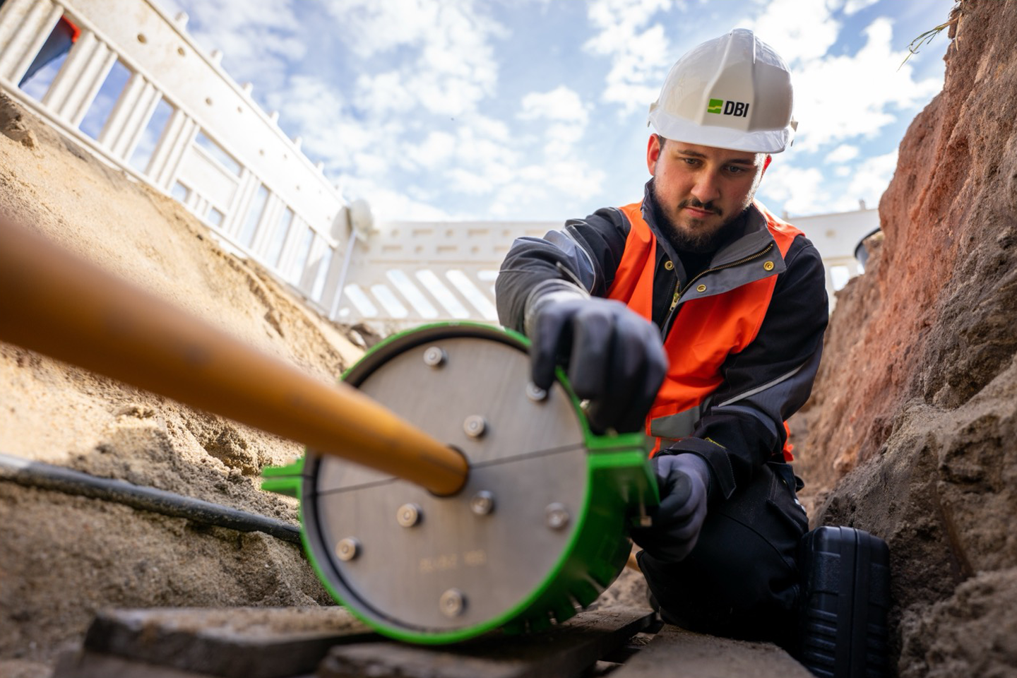 A scientist is sitting in a trench, wearing a helmet and working on a thin tube to which a round measuring device is attached