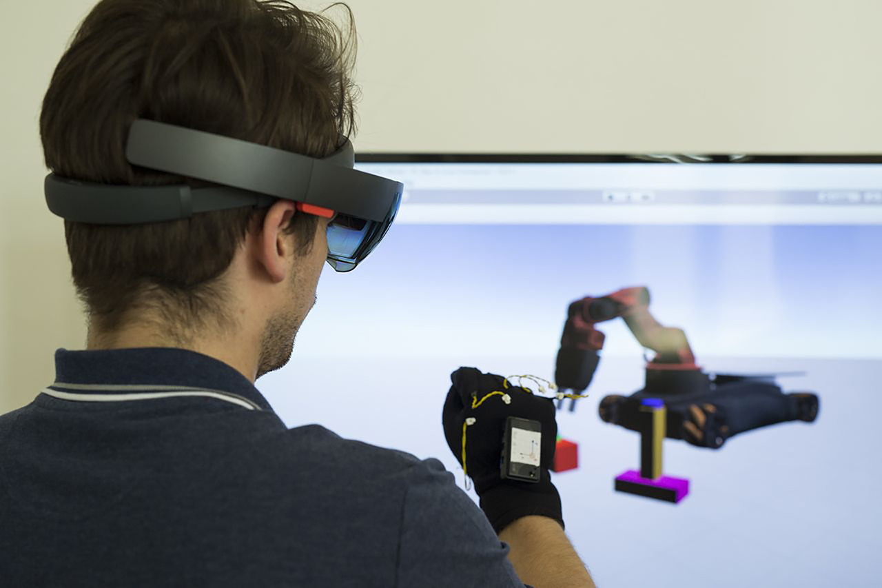 A person wearing VR goggles and a VR glove in front of a monitor; the monitor displays a model of an industrial robot arm to be controlled; clicking the link takes you to the project website