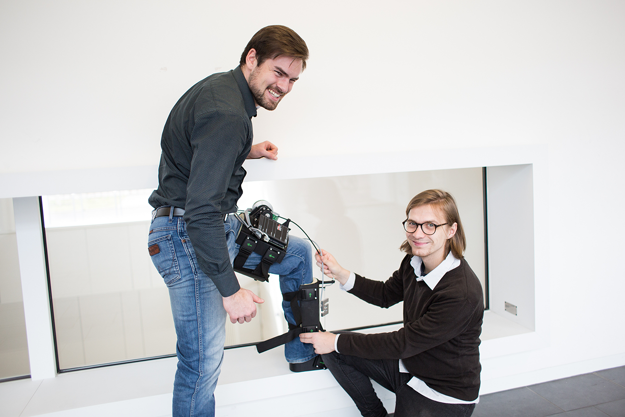 Exoskeleton presentation: A man is wearing an exoskeleton on his leg, which is elevated; other scientist presents the design; the link leads to the project website