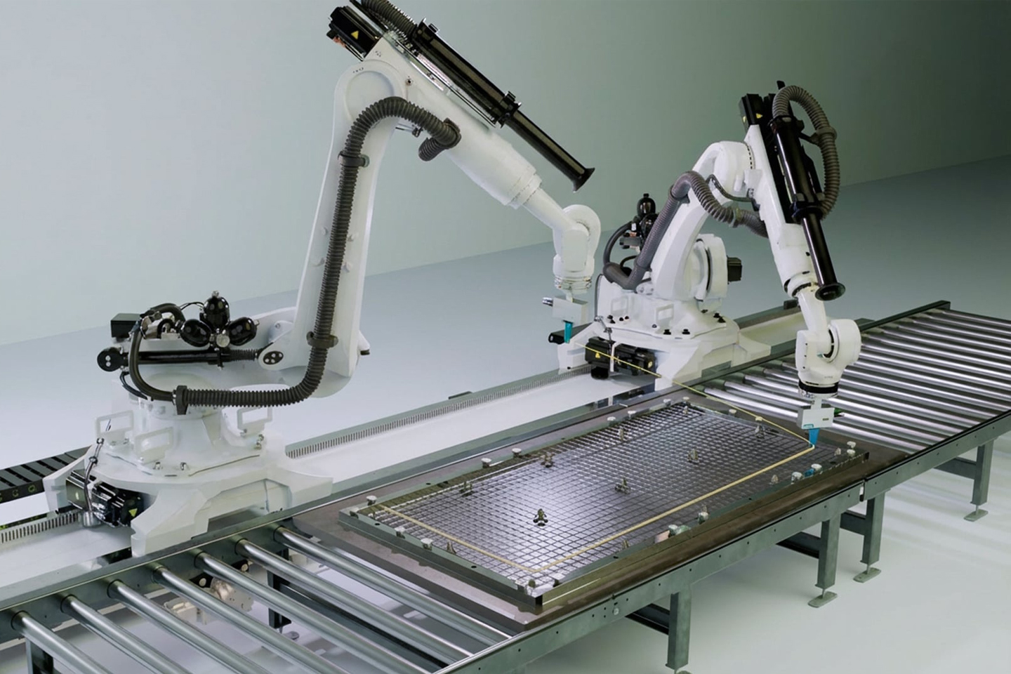 Two white Kuka robotic arms, mounted on a rail, move along a piece of carbon-reinforced concrete and install wires for the manufacture of the product.