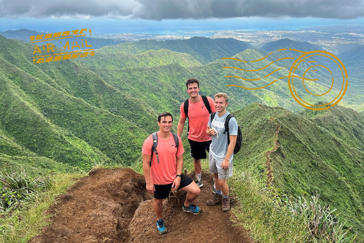 Foto von drei Studierenden, die in Kamera schauen und auf dem Gipfel eines Berges auf Hawaii mit Blick nach Honolulu/Verlinkung führt zum Auslandsbericht